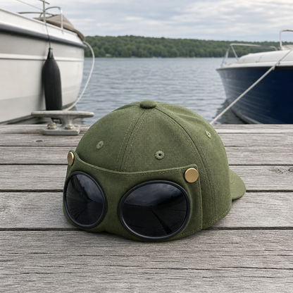 An olive green aviator-style cap with large round black sunglasses attached on a weathered wooden dock, with boats and water in the background.