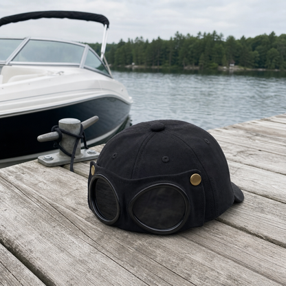 A black vintage-style aviator cap with round dark lenses and brass rivets sits on a weathered wooden dock, with a white boat and a tree-lined lake in the background.